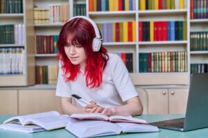 Meisje met roodgeverfd haar, laptop, koptelefoon en boeken in schoolbibliotheek
