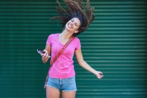lachende vrouw met roze shirt, oortjes in en telefoon in haar hand zwiept met haar lange haar